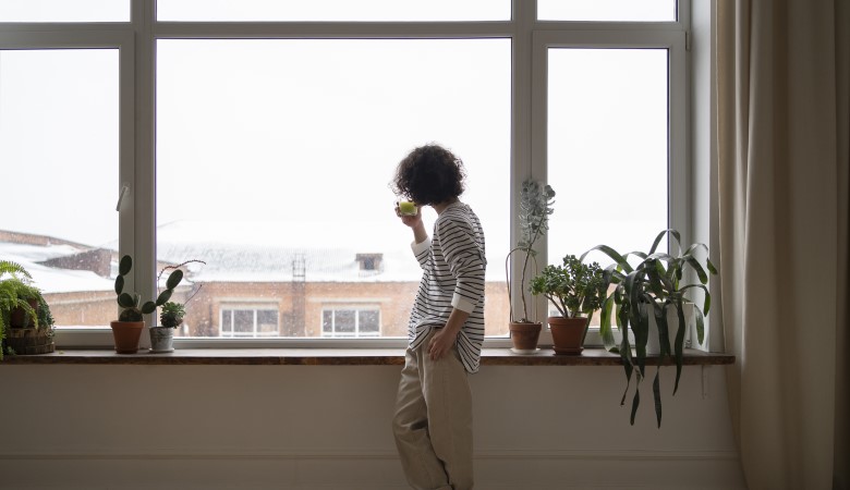 Une personne debout, face à de grandes fenêtres, regardant le paysage en buvant un thé