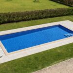 Piscine creusée dans un jardin, sur une pelouse entretenue. Le tour de la piscine est carrelé et il y a deux transats.