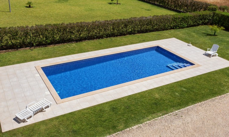 Piscine creusée dans un jardin, sur une pelouse entretenue. Le tour de la piscine est carrelé et il y a deux transats.