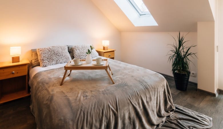 Petite chambre mansardée dans une maison avec un petit déjeuner sur le lit. Le lit dispose également d'une couverture moelleuse et de deux oreillers moelleux. Sur les côtés, il y a des tables de chevet avec des lampes allumées.