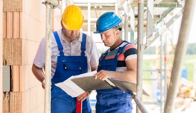 Deux hommes, en tenue de travail, en train de lire un plan à côté d'une maison en chantier