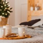 Focus sur un plateau en bois avec des tasses posé sur un lit. Au fond, on voit une guirlande et un petit sapin de Noël.