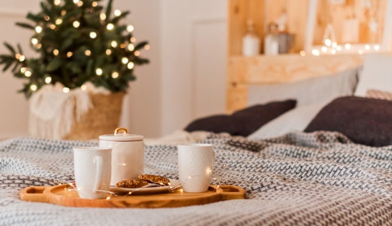 Focus sur un plateau en bois avec des tasses posé sur un lit. Au fond, on voit une guirlande et un petit sapin de Noël.