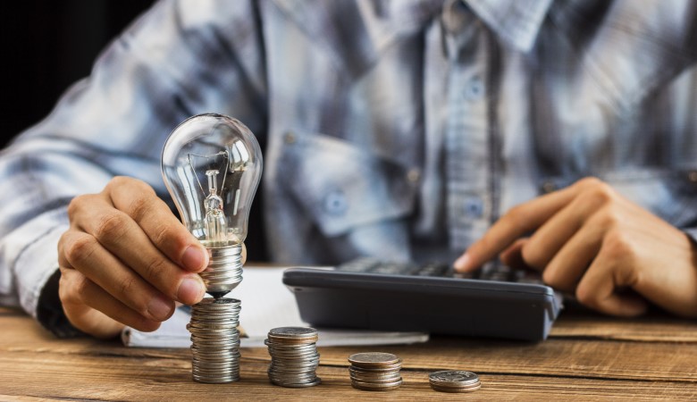 Main d'un homme qui tient une ampoule entre ses doigts et qui utilse une calculatrice de l'autre main