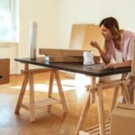 une femme en train de manger et de consulter sa tablette sur une table. La table est dans une pièce vide entourée de carton.