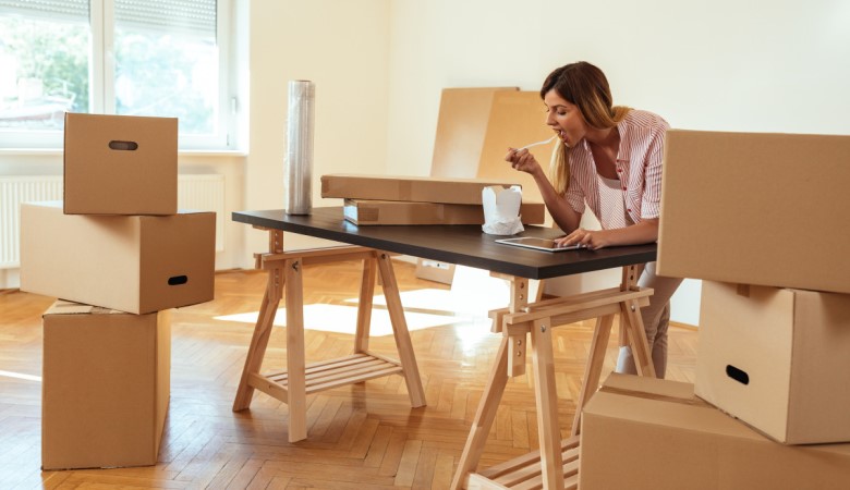 une femme en train de manger et de consulter sa tablette sur une table. La table est dans une pièce vide entourée de carton.