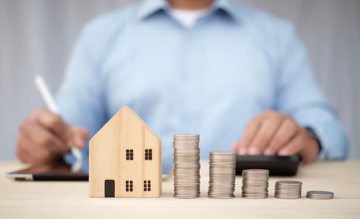 En premier plan, il y a une maison en bois avec des pièces d'argent. En second plan, il y a un homme qui travaille sur son bureau.