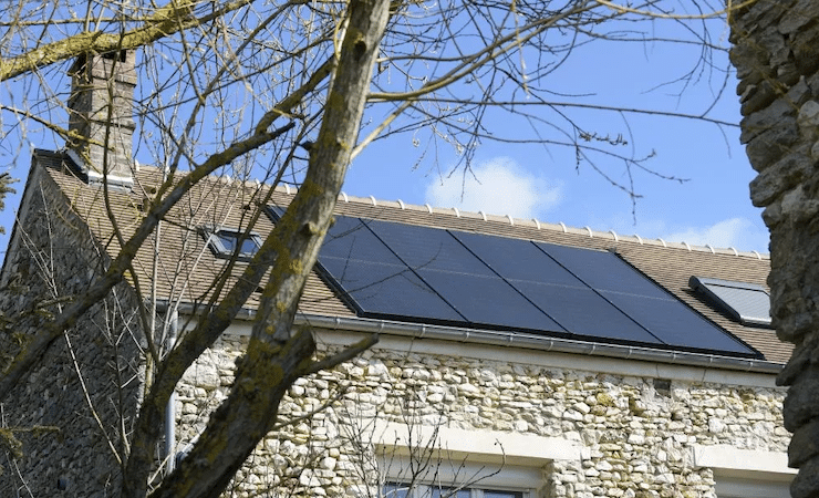 En premier plan, des branches d'arbres. En second plan, des panneaux photovoltaïques sur une toiture.