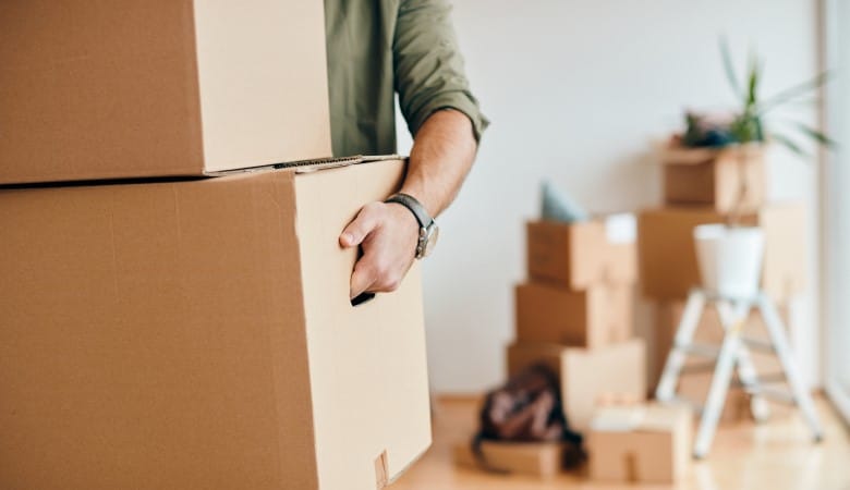 homme qui tient deux cartons dans ses bras. Au fond on aperçoit une pièce vide avec une pile de carton remplis et fermés.