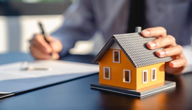 Une maison fictive représentant la vente d'une maison. En arrière-plan, un monsieur signant un contrat.