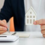 Une personne tenant une maison fictibe dans une main et un stylo de l'autre, à côté se trouve une calculatrice et un papier.