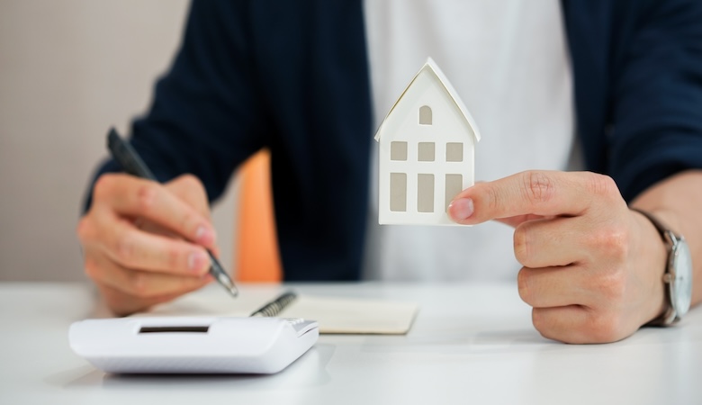 Une personne tenant une maison fictibe dans une main et un stylo de l'autre, à côté se trouve une calculatrice et un papier.