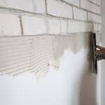 Application de colle à carrelage sur un mur blanc avec une truelle dentelée, prêt pour poser des carreaux effet brique