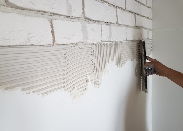 Application de colle à carrelage sur un mur blanc avec une truelle dentelée, prêt pour poser des carreaux effet brique