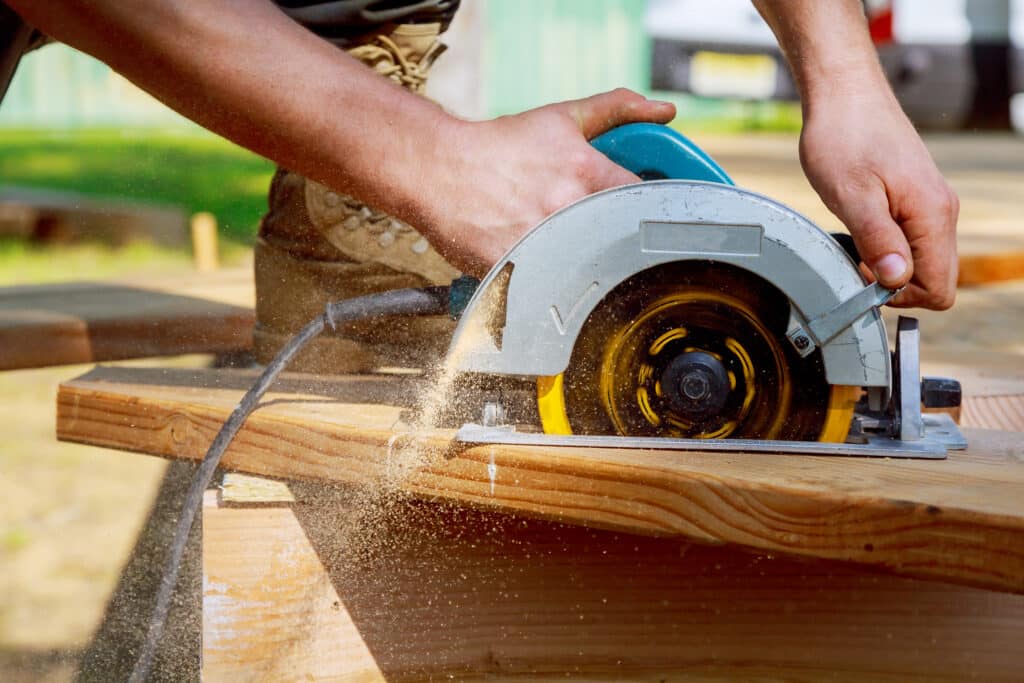 Homme utilisant une scie circulaire Racetools électrique pour couper une planche de bois, avec de la sciure qui s'échappe de la lame.