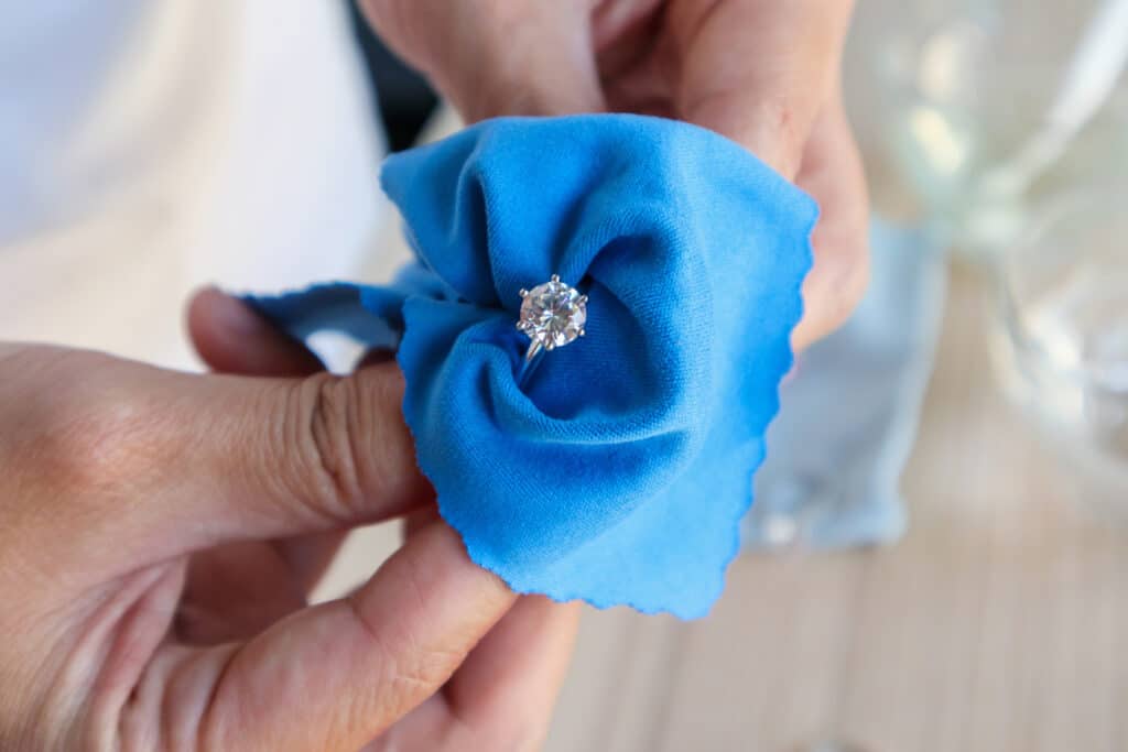 Main tenant une bague en or sertie d’un diamant, nettoyée avec un chiffon doux en microfibre bleu.